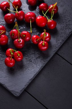 Kayrak tahtasında kiraz. Karanlık bir arka planda tatlı kirazlar. Siyah tahtalarda su damlalarında kırmızı meyveler. Sağlıklı yiyecekler. Üst te. Kopyalama alanı