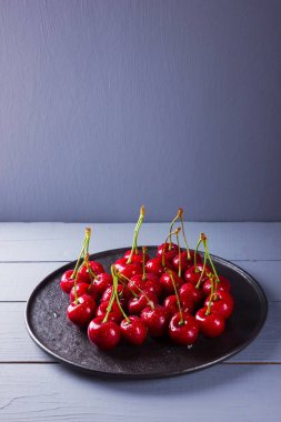 Gri bir arka planda kiraz. Siyah tabakta kırmızı kirazlar. Ahşap tahtalarda su damlalarında çilek. Sağlıklı yiyecekler. Kopyalama alanı