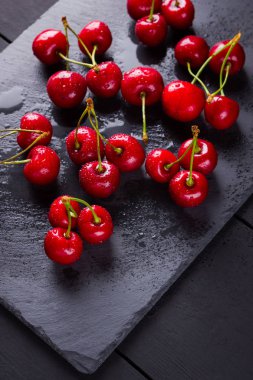 Kayrak tahtasında kiraz. Karanlık bir arka planda tatlı kirazlar. Siyah tahtalarda su damlalarında kırmızı meyveler. Sağlıklı yiyecekler. Üst te. Kopyalama alanı