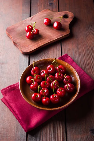 Cherries on a clay plate. Sweet cherries on a wooden board. Red berries in drops of water on a red napkin. Rustic style. Copy space