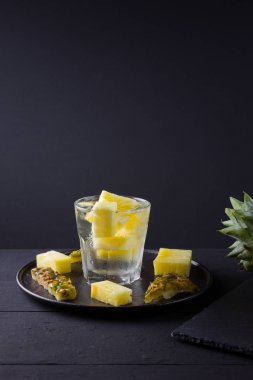 Koyu renkli bir arka planda ananas. Siyah bir tabakta dilimlenmiş ananas. Buz ve ananas dilimleri ile içilir. Aşılanmış su. Kopyalama alanı
