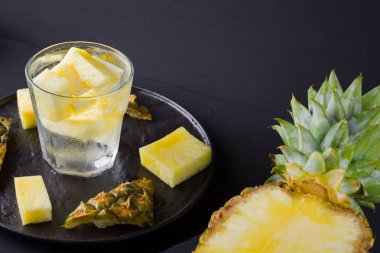 Koyu renkli bir arka planda ananas. Siyah bir tabakta dilimlenmiş ananas. Buz ve ananas dilimleri ile içilir. Aşılanmış su. Kopyalama alanı