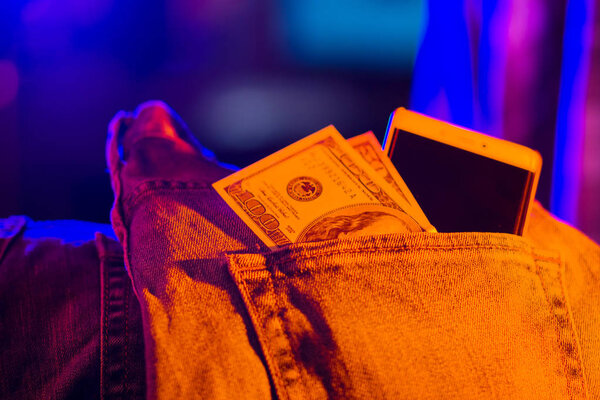 Сто долларов купюры и смартфона в кармане синих джинсов, тонированные. Доллары в неоновом свете. Банкноты 100 и 50 американских долларов и мобильный телефон в джинсовом кармане

