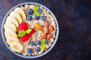 Sağlıklı kahvaltı. Yabanmersinli yulaf ezmesi, muz ve ahududu. İçinde meyve ve fındık olan yulaf ezmesi. Koyu arkaplanda pişmiş yulaf ezmesi. Vejetaryen yemeği.