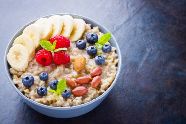Sağlıklı kahvaltı. Yabanmersinli yulaf ezmesi, muz ve ahududu. İçinde meyve ve fındık olan yulaf ezmesi. Koyu arkaplanda pişmiş yulaf ezmesi. Vejetaryen yemeği.