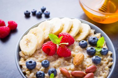 Sağlıklı kahvaltı. Meyveli, fındıklı ve ballı yulaf ezmesi. Yabanmersinli yulaf ezmesi, muz ve ahududu. Koyu arkaplanda pişmiş yulaf ezmesi. Vejetaryen yemeği.