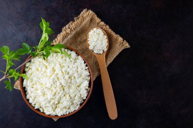 Koyu bir arka planda süzme peynir ve yeşil yapraklar. Siyah arka planda taze süzme peynir ve ahşap kaşık. Çuval bezinde yumuşak peynir. Üst Manzara. Boşluğu kopyala