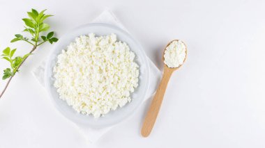 Beyaz arka planda beyaz tabakta süzme peynir. Yumuşak peynir ve tahta kaşık. Beyaz peçetede yeşil yapraklı süzme peynir ve dal. Üst Manzara. Boşluğu kopyala