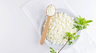Beyaz arka planda beyaz tabakta süzme peynir. Yumuşak peynir ve tahta kaşık. Beyaz peçetede yeşil yapraklı süzme peynir ve dal. Üst Manzara. Boşluğu kopyala