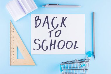 Okul konseptine geri dönelim. Alışveriş arabası ve mavi arka planda kırtasiye malzemeleri. Okul için kalem, üçgen ve not defteri. Ders çalışma zamanı. Üst görünüm