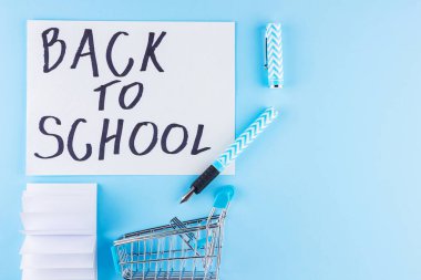 Okul konseptine geri dönelim. Alışveriş arabası ve mavi arka planda kırtasiye malzemeleri. Okul için kalem, kalem ve not defteri. Üst görünüm