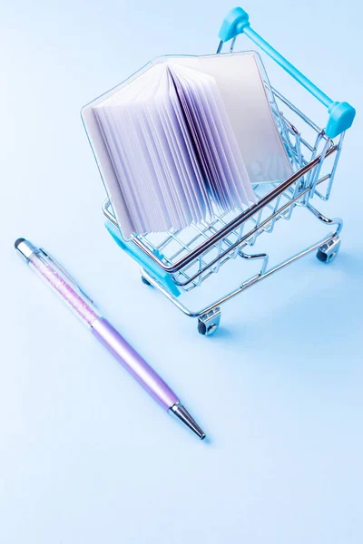 Shopping cart with school supply. School notepad with holographic cover. Notepad and pen in shopping trolley. Back to school concept