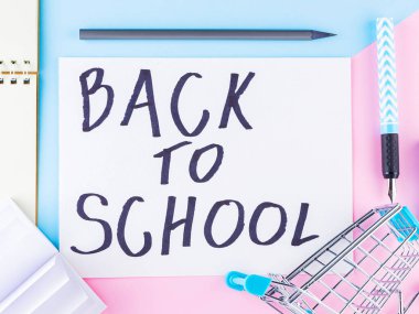 Okul konseptine geri dönelim. Pembe mavi arka planda alışveriş arabası ve kırtasiye malzemeleri. Kalem, not defteri, kalem ve alışveriş arabası. Ders çalışma zamanı. Üst görünüm