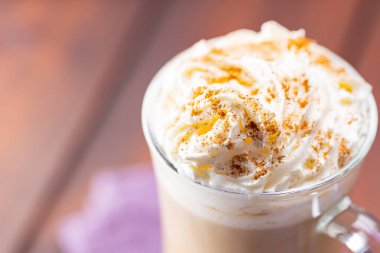 Cam bardakta balkabağı aromalı latte. Tahta bir tahtanın üzerinde krem şantili ve baharatlı balkabaklı latte. Keten peçetede sıcak sonbahar kahvesi.