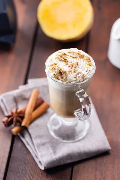 Pumpkin latte, milk jug and a piece of pumpkin. Pumpkin latte with whipped cream and spices on a wooden boards. Hot autumn coffee drink on a linen napkin