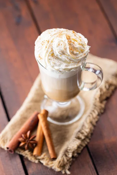 Pumpkin spice latte in a glass mug. Pumpkin latte with whipped cream and spices on a wooden background. Hot autumn coffee drink on a burlap