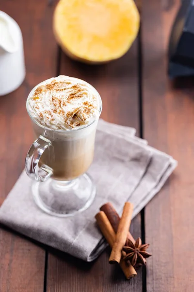 Pumpkin latte, milk jug and a piece of pumpkin. Pumpkin latte with whipped cream and spices on a wooden boards. Hot autumn coffee drink on a linen napkin