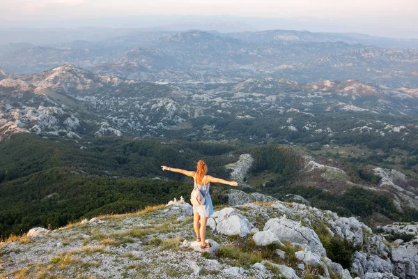 Hermosa chica turística se encuentra en la cima de un acantilado y admira el hermoso paisaje de la cordillera en Montenegro. Concepto de turismo, viajes y vacaciones de verano — Foto de Stock