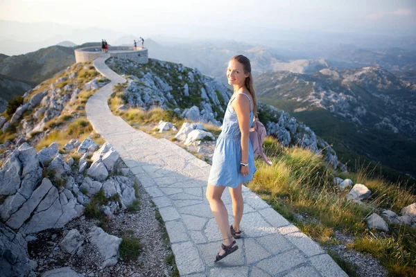 Hermosa chica turística se encuentra en la cima de un acantilado y admira el hermoso paisaje de la cordillera en Montenegro. Concepto de turismo, viajes y vacaciones de verano — Foto de Stock