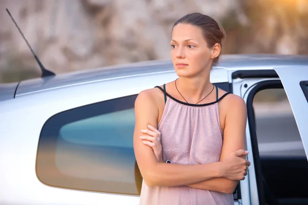 Young sad woman standing near the road by the broken car in the middle of nowhere. Waiting car tow service.