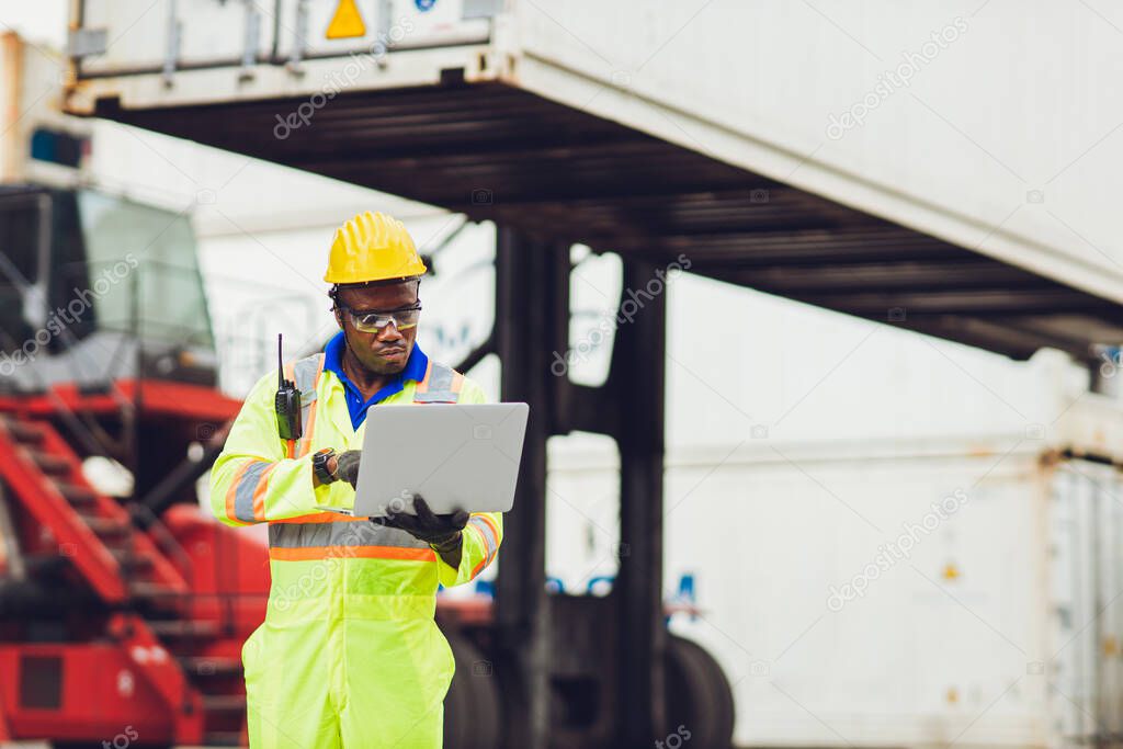 Black African worker working in logistic shipping using laptop to control loading containers at port cargo for import export goods foreman looking high for future