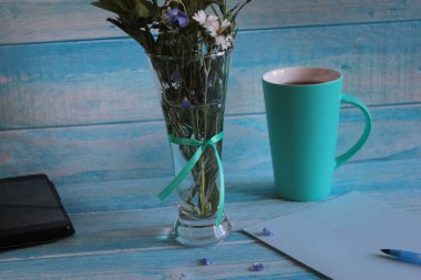 Çay, telefon, yabani çiçek buketi ve mavi ahşap arka plan üzerinde kağıt boş bir levha ile Kupa.