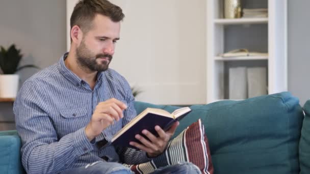 Junger Mann liest Buch, sitzt auf Sofa — Stockvideo