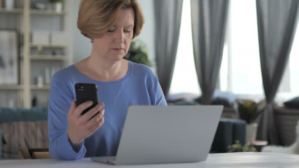 Стара старша жінка використовує смартфон і ноутбук на робочому місці — стокове відео