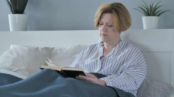 Стара старша жінка читає книгу сидячи в ліжку — стокове відео