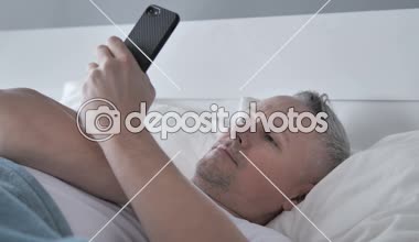 Gri saç adam kullanma Smartphone Lying yatakta iken