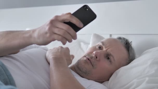 Hombre Pelo Gris Reaccionando Pérdida Mientras Usa Teléfono Inteligente Cama — Vídeo de stock