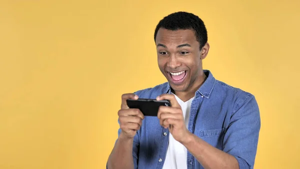 在黄色背景中隔离的智能手机上玩游戏的非洲青年男子 — 图库照片