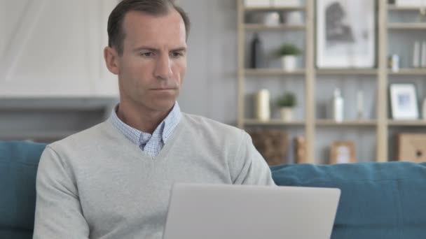 Hombre de mediana edad trabajando en el ordenador portátil mientras está sentado en el sofá — Vídeos de Stock