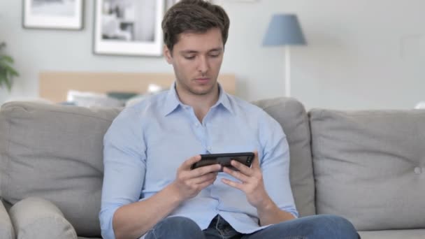 Joven guapo animando el éxito en el teléfono inteligente mientras está sentado en el sofá — Vídeo de stock