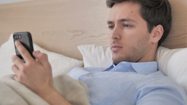 Jóvágású fáradt fiatal ember használ smartphone-ban ágy — Stock videók