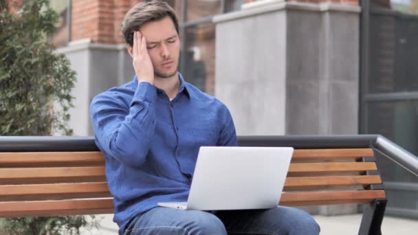 Сидящий на открытом воздухе молодой человек с головной болью с помощью ноутбука — стоковое видео