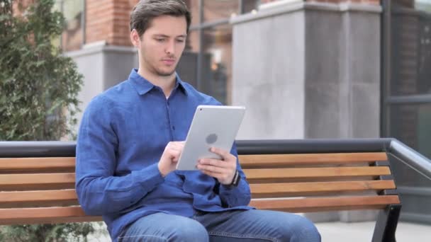 Młody człowiek za pomocą tabletu, siedząc na ławce — Wideo stockowe