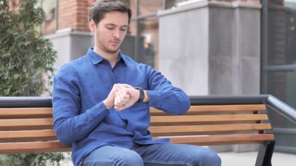 Νέος άνθρωπος χρησιμοποιώντας έξυπνο ρολόι, υπαίθρια κάθεται στο πάγκο — Αρχείο Βίντεο