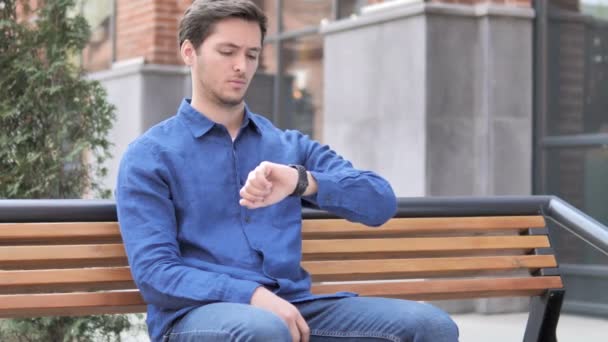 Περιμένοντας νέος άνθρωπος βλέποντας τον χρόνο στο ρολόι, καθισμένος στον πάγκο — Αρχείο Βίντεο