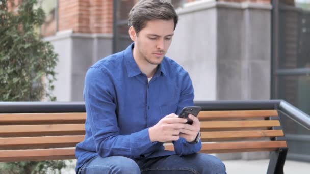 Ung man skriva meddelande på smartphone, sittande utomhus på bänken — Stockvideo