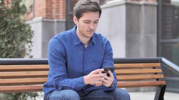 Jonge man viert Win op smartphone, zittend buiten op Bank — Stockvideo
