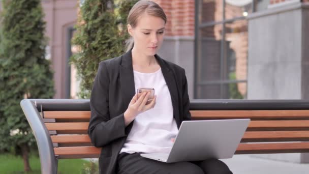 Ung affärskvinna med hjälp av smartphone och laptop, sitter på bänken — Stockvideo