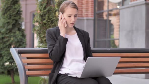 Молодая предпринимательница разговаривает по телефону, сидит на скамейке запасных — стоковое видео