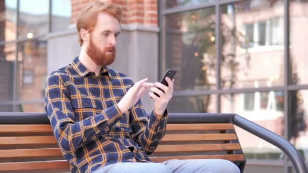 Redhead skägg ung man firar vinna på smartphone, sittande utomhus på bänken — Stockvideo