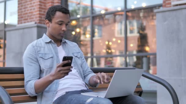 Afrikansk man med hjälp av smartphone och laptop, sitter på bänken — Stockvideo