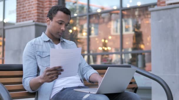 Африканський чоловік читає контракт і працює на ноутбуці, сидячи на лавці — стокове відео