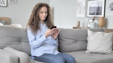 Kanepe üzerinde rahatlatıcı iken Smartphone kullanarak genç kıvırcık saç kadın