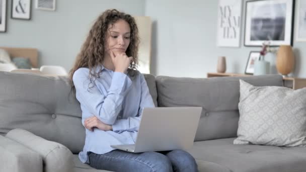 Mujer de pelo rizado pensando y trabajando en el ordenador portátil mientras está sentado en el sofá — Vídeo de stock