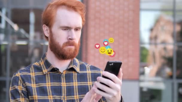 Redhead baard man met behulp van smartphone, vliegende emojis, smileys en likes — Stockvideo