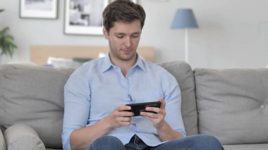Genç adam oyun oyunları Smartphone, kanepede oturan